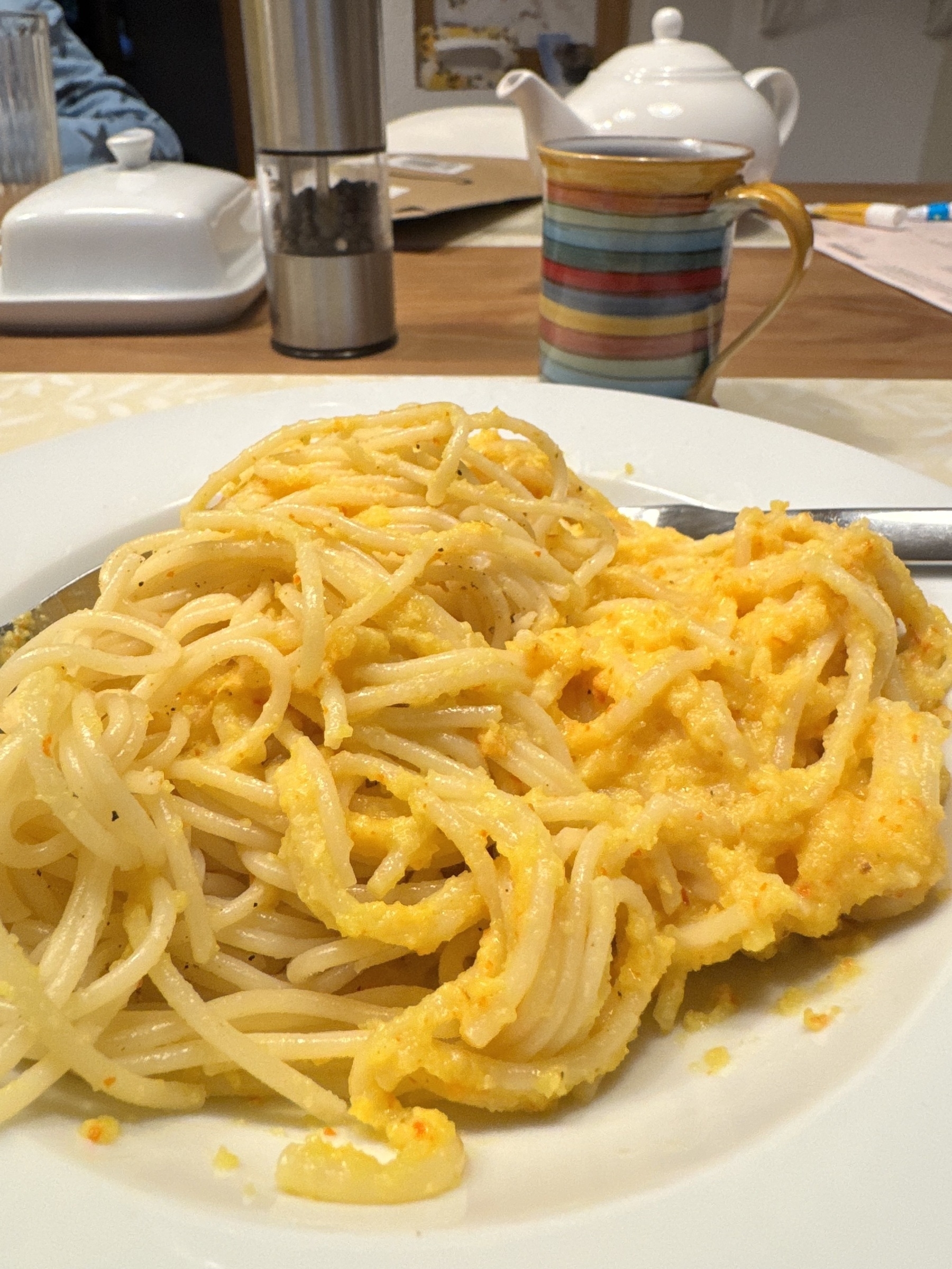 Teller mit Spaghetti in cremiger gelber Sauce, im Hintergrund bunte gestreifte Tasse und weiße Teekanne