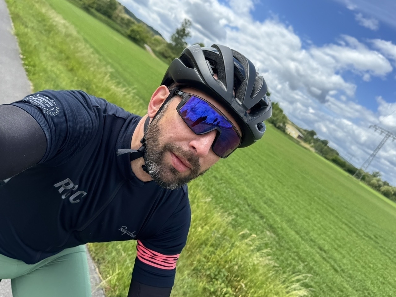 Auto-generated description: Ein Radfahrer mit Helm und Sonnenbrille fährt auf einer ländlichen Straße neben einem grünen Feld unter einem bewölkten Himmel.