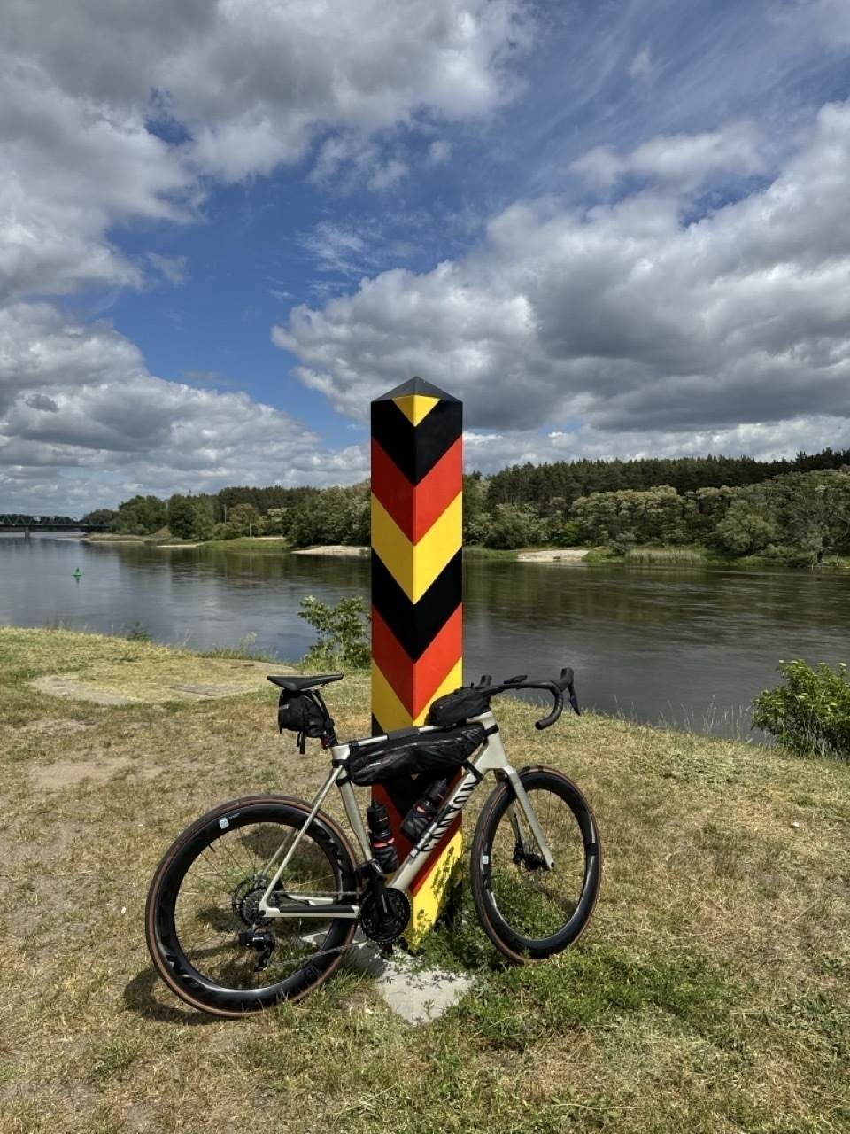 Auto-generated description: Ein Fahrrad lehnt an einem Grenzpfosten in den Farben Schwarz, Rot und Gold, mit Fluss und Landschaft im Hintergrund.